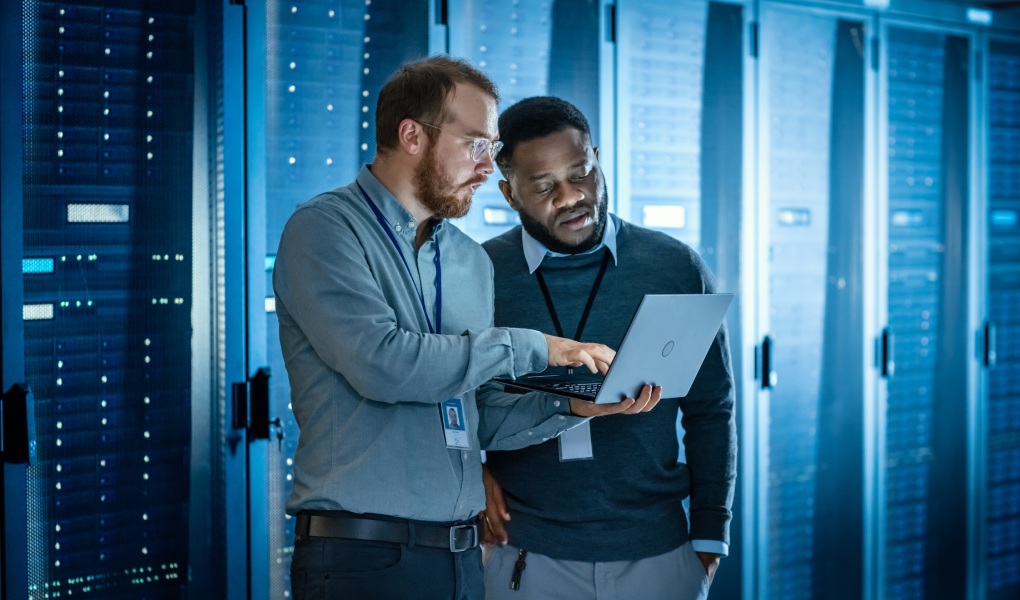 Bearded IT Technician in Glasses with Laptop Computer and Black Male Engineer Colleague are Using Laptop in Data Center while Working Next to Server Racks Running Diagnostics or Doing Maintenance Work