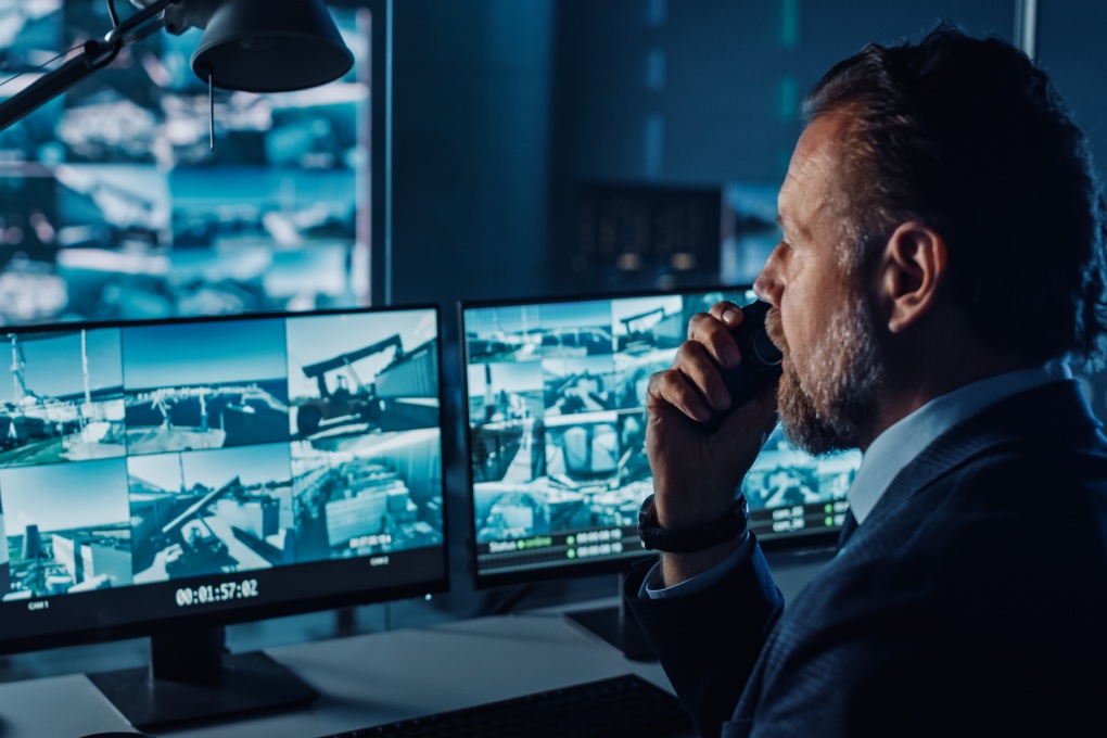 Male Officer Works on a Computer with Surveillance CCTV Video in a Harbour Monitoring Center with Multiple Cameras on a Big Digital Screen. Employee Uses Radio to Give an Order or Report.