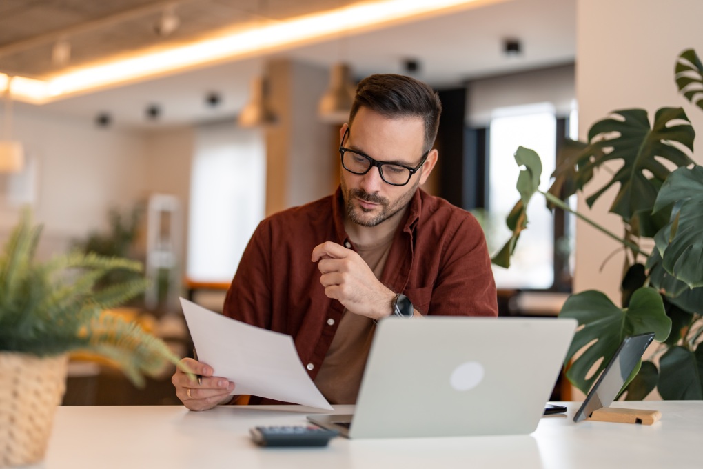 Serious millennial man using laptop sitting at the table in a home office, focused guy looking at the paper, communicating online, writing emails, distantly working or studying on computer at home.