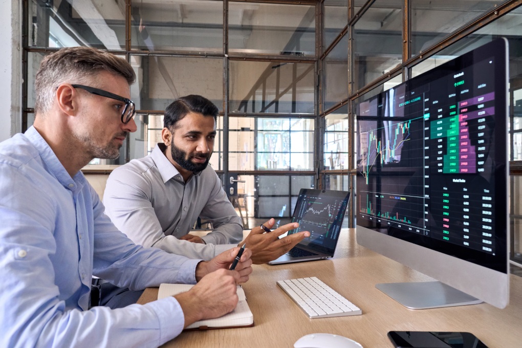 Two diverse crypto traders brokers stock exchange market investors discussing trading charts research reports growth using pc computer looking at screen analyzing invest strategy, financial risks.