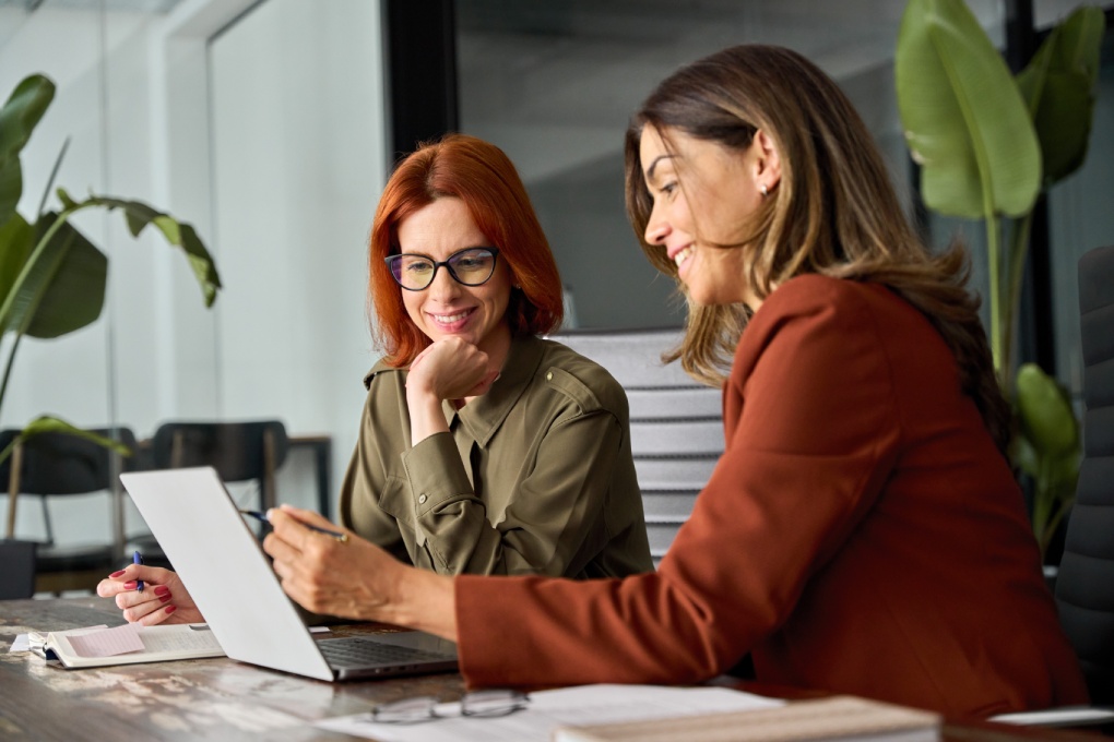 Two happy busy female employees working together using computer planning project. Middle aged professional business woman consulting teaching young employee looking at laptop sitting at desk in office