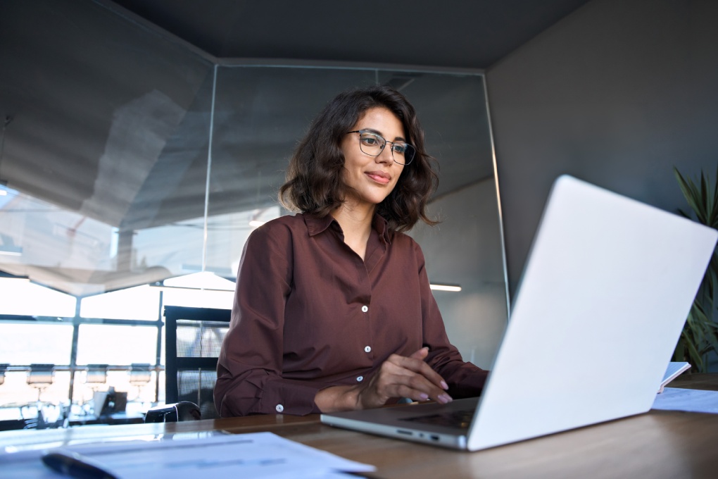 Young professional it specialist latin hispanic business lady working on laptop pc sitting at desk in modern office space. 30s middle eastern indian woman using computer technology app for work online