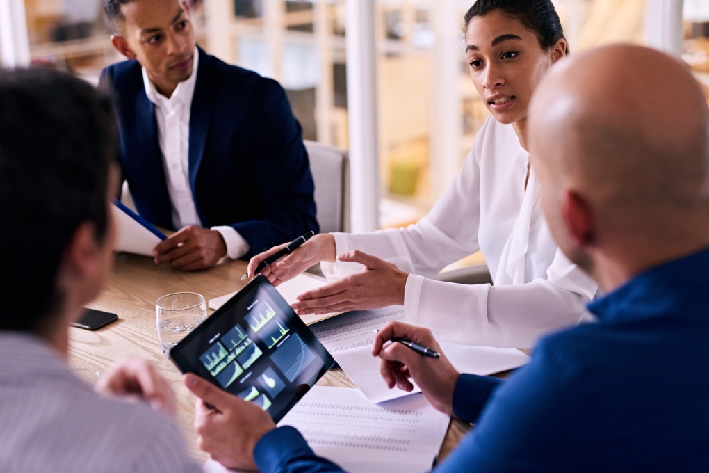 Business meeting between four upper management board members in the new modern office conference room with technology integrated in the form of an electronic tablet.