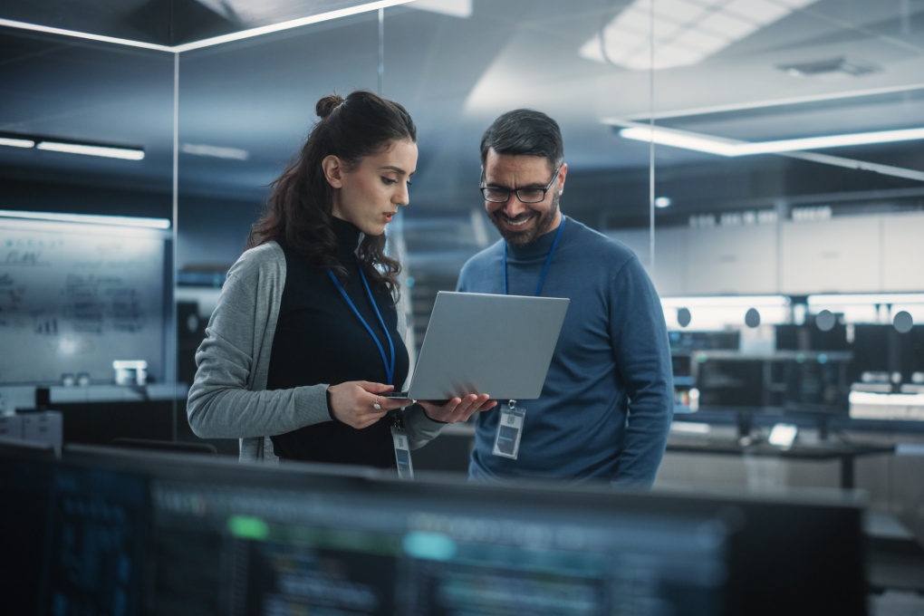 Portrait of Two Happy Female and Male Engineers Using Laptop Computer to Analyze and Discuss How to Proceed with the Artificial Intelligence Software. Casually Chatting in High Tech Research Office